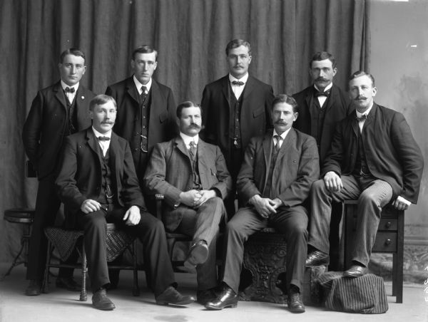 Eight Scandinavian men in suits and ties sit and stand in two rows posing for a formal studio portrait. Behind them is a curtain and part of a painted backdrop.