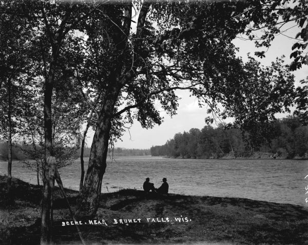 Two men sit on the shore of the Chippewa River near Brunet Falls. The light is dim. There are large trees behind them.
