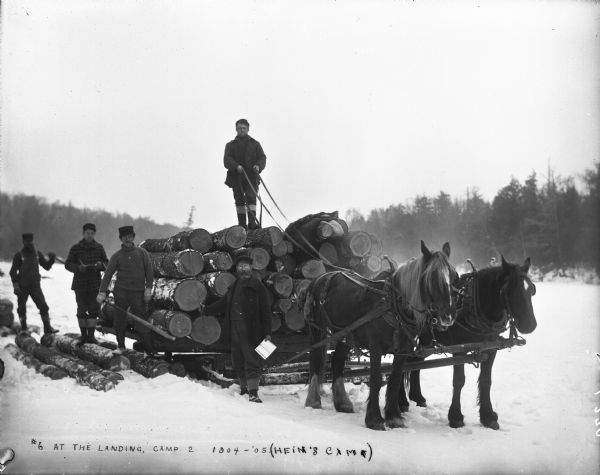 Five men stand on or nearby a sled loaded with logs and pulled by two horses at the landing at Hein's logging camp. There is snow on the ground.