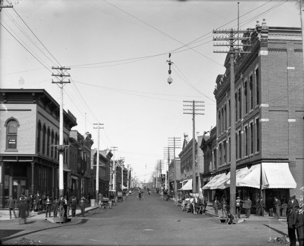 View down middle of Bridge Street, looking north. A busy retail street with buildings on both sides, numerous horses-and-buggies, and many pedestrians on the sidewalks. A street light hangs over the intersection.
