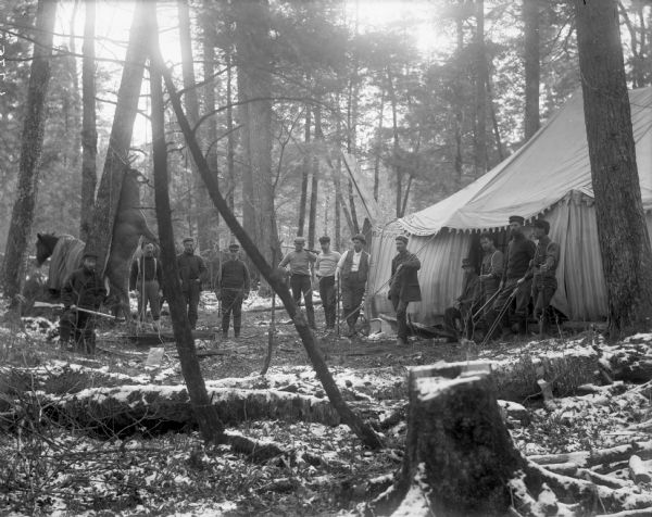 About a dozen men stand in the woods by a large tent. The carcass of a deer is hanging from one of the trees. A horse stands nearby. There are some traces of snow on the ground.