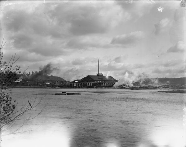 The Big Lumber Mill seen from across the Chippewa River under cloudy skies, with smoke coming out of several smokestacks