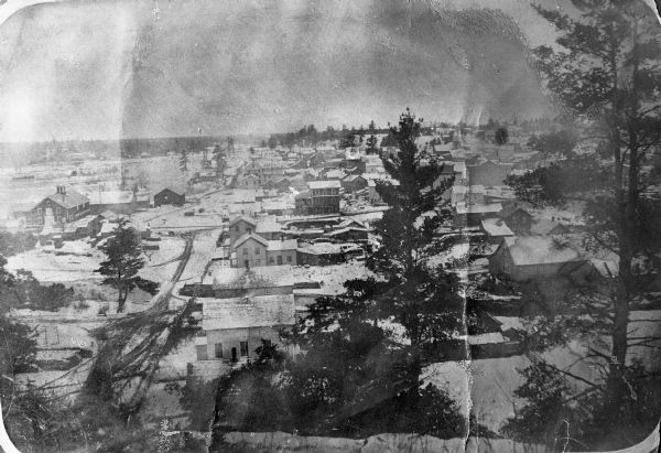 Elevated copyprint view of Eau Claire showing numerous houses and commercial buildings, with snow on the ground and large pine trees in the foreground
