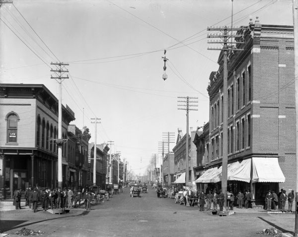 Bridge Street, looking north, with commercial buildings lining both sides of the street, and numerous horses and buggies on the street, and many people standing on the sidewalks and street corners.