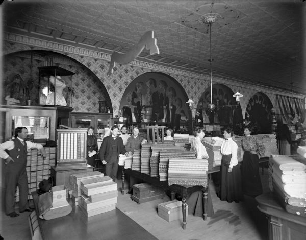 Several men and women standing inside a department store next to tables piled with bolts of fabric. The store has a pressed tin ceiling.