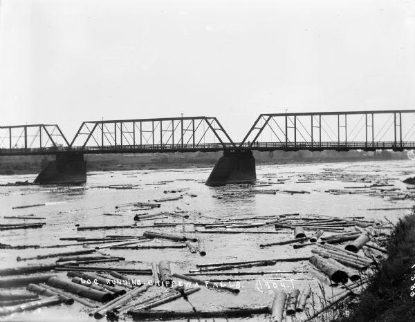 Logs floating down the river beneath the bridge on the Chippewa River. On the far shoreline are billboards. One advertises for "J.R. Sharp Hardware." Men are standing on the bridge.