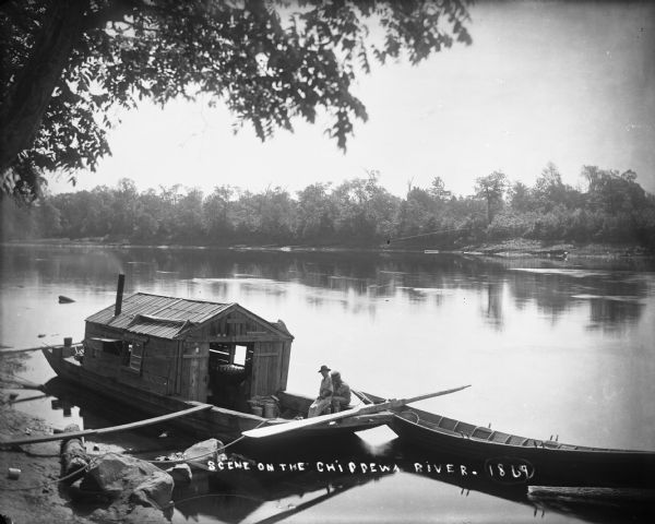 Two men, one wearing an apron, sit in a wanigan tied to the shore of the Chippewa River near Chippewa Falls. The inside of the boat appears to have a pile of clams on a box near a window. Two clam shells wedged above the doorway support the notion that the items on the box are clams. There is also a horse shoe hanging above the door. There is a bateau tied to the wanigan.