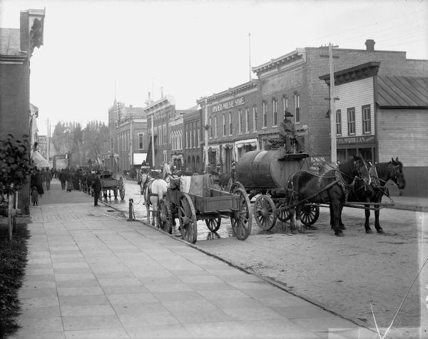 Horse-drawn wagons, including a water wagon, stand on a street next to a broad sidewalk in a commercial district. The storefronts of P.L. McQuillan and Consumer's Wholesale Store are on the far side of the street. There are numerous pedestrians on the sidewalk. There is a steep hill in the background.