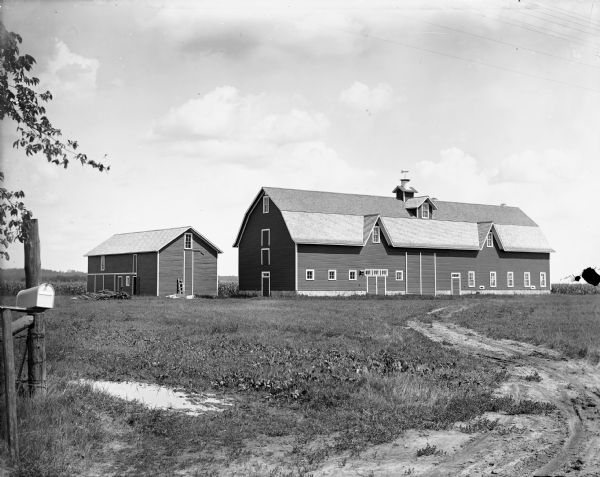 A barn and outbuilding on the Joe Kelley farm in a field at the end of a dirt track. There is a mailbox attached to a post in the left foreground.