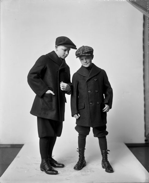 Two young boys in watch caps, heavy wool coats, knickers and boots pose for an advertisement for C-F Woolen Mills in front of a studio backdrop.