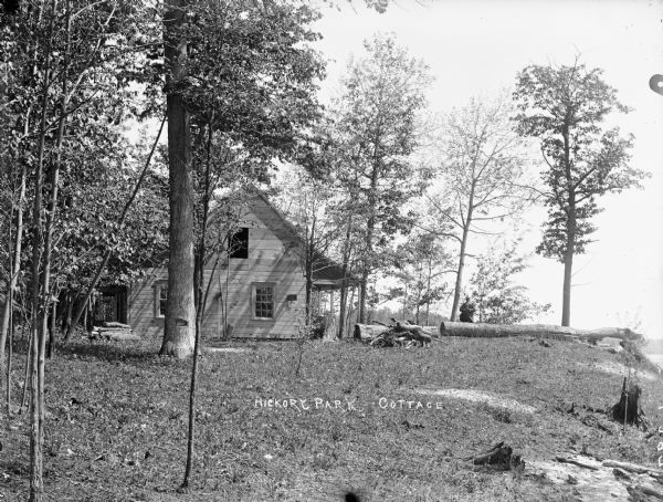 The clapboard Hickory Park Cottage stands at the edge of a wooded area. A man holding a gun stands behind cut logs to the right of the cottage. There appears to be a lake on the far right.