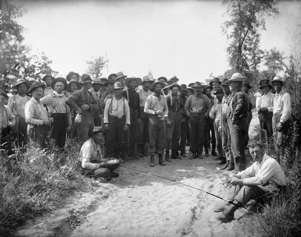 A few dozen men stand on and around a sandy path surrounded by low shrubbery. They are wearing work clothes, and some of them are carrying bowls filled with food.