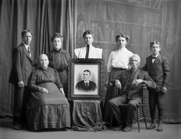 Seven members of the Fragon family sitting and standing for a portrait around a framed painting of a young man, possibly a deceased family member. An older man and woman are seated in the front row and five younger men and women stand in the rear. There is a painted backdrop behind them.