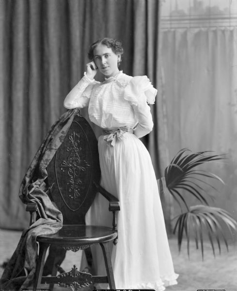 Full-length studio portrait in front of draperies and a painted backdrop of a young woman wearing a full-length white dress with a lacy bodice and a pastel sash standing by a wooden chair with an oval, elaborately carved back, draped with a patterned piece of fabric. A potted fern stands behind her to the right.