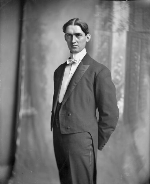 Three-quarter length studio portrait in front of draperies of a man standing in a tuxedo and white shirt and tie, with his hands crossed behind his back.