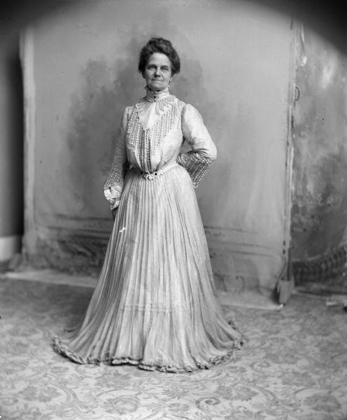 Full-length studio portrait in front of a backdrop of a woman wearing an elaborate light-colored dress standing with one hand behind her back on a patterned rug.