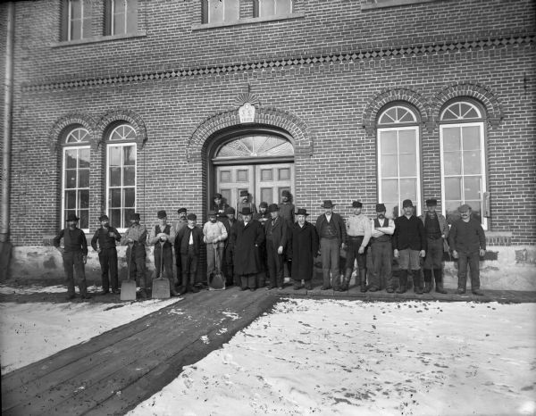 A group of about two dozen male city employees, three carrying shovels, stand in front of the double doors of a large brick building. There is a wooden walkway leading up to the doors and snow is on the ground.