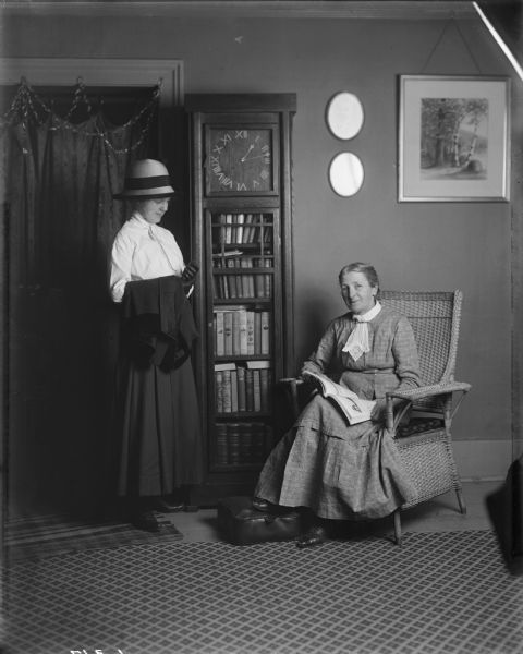 An older female member of the Bish family sits in a wicker chair holding a magazine or book open on her lap. Standing on the left in front of a doorway draped with a curtain, is a younger woman in a dark skirt and white blouse with a dark jacket draped over her right arm. Between them, standing against a wall, is a tall wooden clock with a glass-doored bookcase underneath it filled with books. Two small mirrors and a landscape illustration hang on the wall above the older woman.