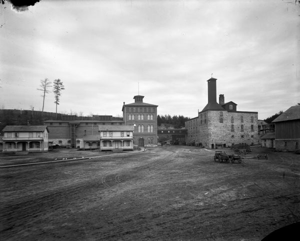 Buildings and grounds of the Leinenkugel Brewery. There are beer wagons in the yard, chickens along a wooden walkway, and men on a horse-drawn wagon near a loading dock.