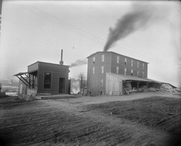 Two buildings on the grounds of the South Side Manufacturing Company stand side-by-side in a furrowed dirt yard. The larger building has a large lean-to filled with lumber. There is a bicycle leaning against the side of the smaller buildings. A plume of smoke is coming from another building somewhere in the background.