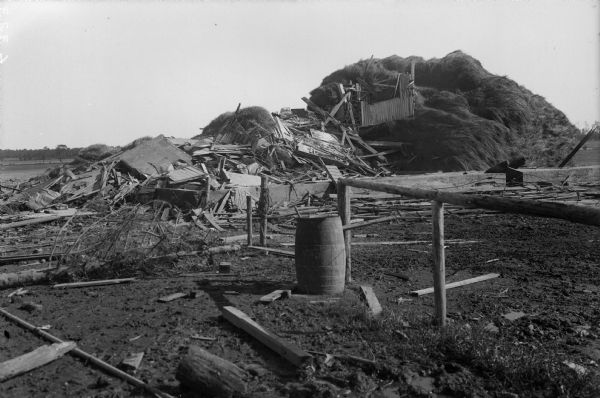 The wreckage of buildings on the John Hedler farm after a tornado. An intact wooden barrel and part of a wooden fence stands in the center foreground. A large pile of hay is visible in the background.