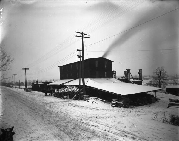 Elevated view from road of a building on the grounds of the South Side Manufacturing Company, surrounded by a lean-to stacked with lumber. Two horses stand hitched to a cart that is backed into the lean-to. There is snow on the ground. On the lower left is a horse.