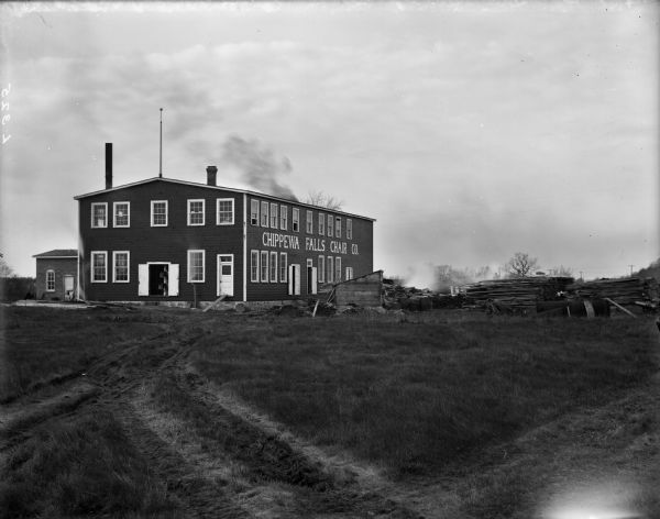 Exterior view of a rectangular clapboard building. On the right side are large letters spelling out: "CHIPPEWA FALLS CHAIR CO." The building stands in a field at the end of a furrowed wagon track in the grass. There is a yard full of lumber on the right.