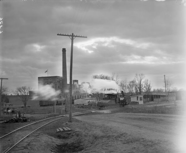 A steam locomotive moves along a set of railroad tracks near a sugar beet processing plant with a tall smokestack. There are several small outbuildings in the vicinity of the plant and along the tracks. A man with a horse-drawn wagon is on the far left.