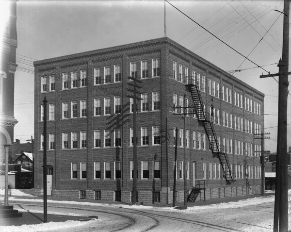 The square, four-story brick Chippewa Shoe Company building, located at the intersection of Bay Street and River Street looking North. There is a fire escape on the right side of the building and a utility pole casts a shadow on the front of the building. Streetcar tracks run down the middle of the street in front of the building. A sign on a small building to the left of the shoe company reads: "Old Virginia Cheroots."