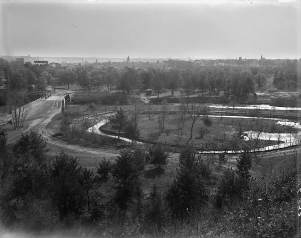 Elevated view of park-like, lightly-wooded area with Duncan Creek running through it. A dirt road runs over a bridge to the left. The skyline of Chippewa Falls is hazy in the distance.
