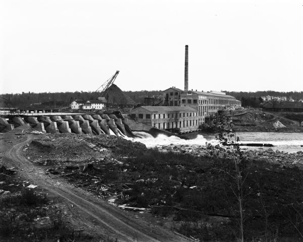 Elevated view across dirt road of a paper mill on the shore of the Chippewa River next to a concrete dam. The mill has a tall smokestack. On the far shoreline are houses.
