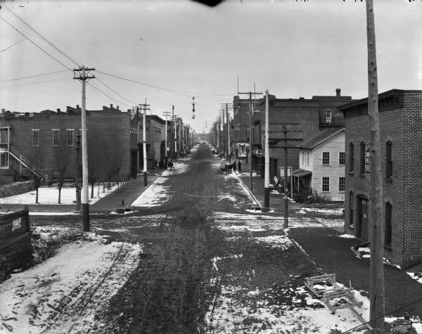 Elevated view looking North down middle of Bridge Street. Both sides of the street are lined with commercial buildings. There are several horse-drawn vehicles standing along the curbs, pedestrians on the sidewalks, and traces of snow on the ground.