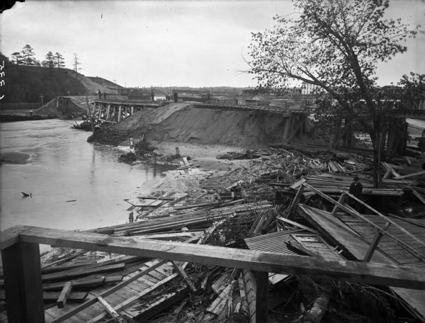 View of the wreckage of wooden structures lying on the shoreline of a river after a flood. A dam across the river has been partially washed away. A man is standing among the debris.