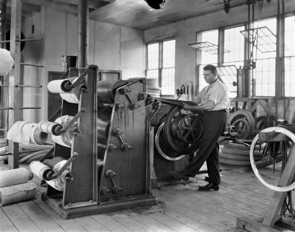 Interior view of the Boone Tire Factory. A man is using a machine to wrap canvas around a rubber tire.