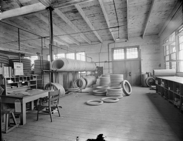 Interior view of room at the Boone Tire Factory. Tires are stacked on the floor and in racks. In the left foreground is a large desk with hutch and two chairs. A chain link fence separates part of the room.