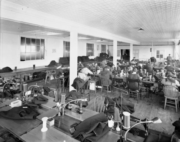 About twenty women sit at tables sewing on sewing machines in the sewing room of the Chippewa Woolen Mill.