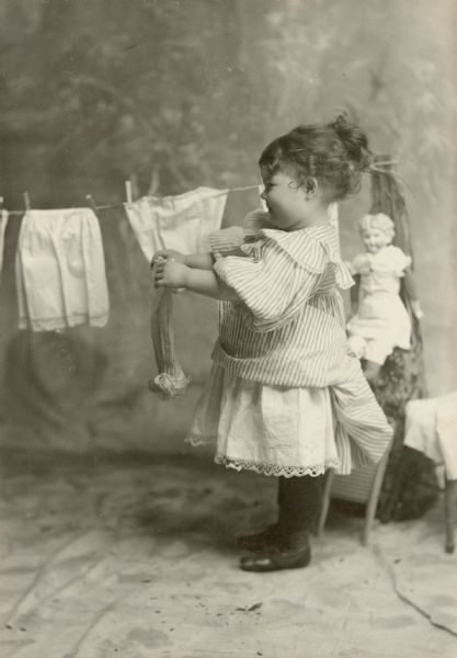 Margery Bish as a little girl. She is wearing a striped dress while hanging clothes on a clothesline. There is a doll behind her on the right.