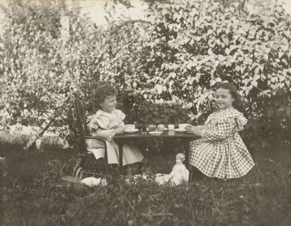 Margery Bish (left) and another little girl, unidentified, sit at a low outdoor table set with a tea service. There is a china doll propped under the table.