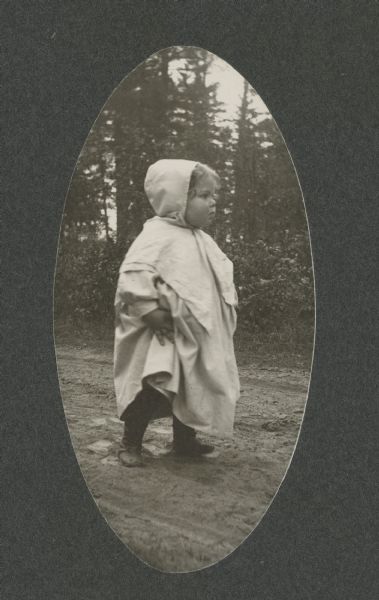 Margery Bish, the toddler daughter of the photographer, stands on a dirt road wearing a hooded cloak.