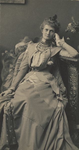Mary Bell, daughter of Mr. Bell, the President of the First National Bank of St. Louis, sits with head on hand and elbow leaning on a carved wooden pedestal that also holds a vase of flowers. A caption by an unknown person indicates that Mary for "a very long time came to our Chippewa Falls house parties. Many interesting people."