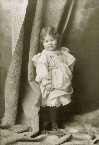 Margery Bish, the young daughter of the photographer, stands in front of a draped curtain.