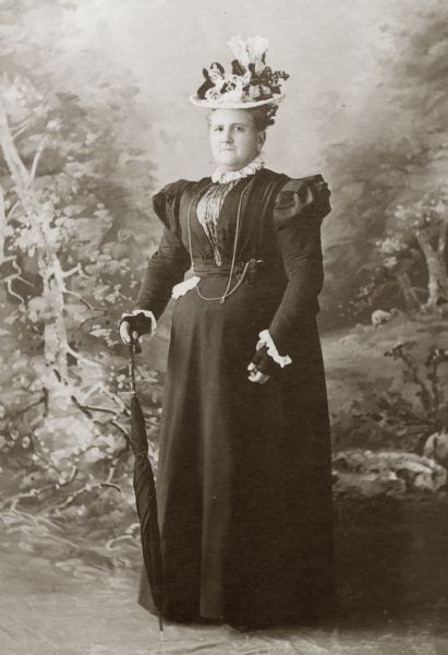 Full-length portrait of a lady in a floor-length dark dress and elaborate flowered hat standing and holding an umbrella in front of a painted studio backdrop of a forest. She wears fingerless gloves and has a long chain around her neck attached to what is presumably a watch in her pocket.