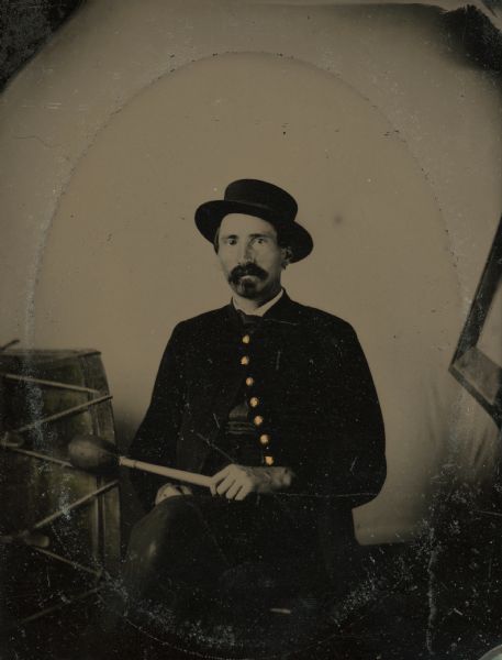 Tintype portrait of Jacob Brant of Brodhead, Wisconsin, member of the 3rd Wisconsin Infantry band. He is seated in uniform next to a bass drum and holds a padded drumstick. The buttons on his uniform are hand-colored gold.