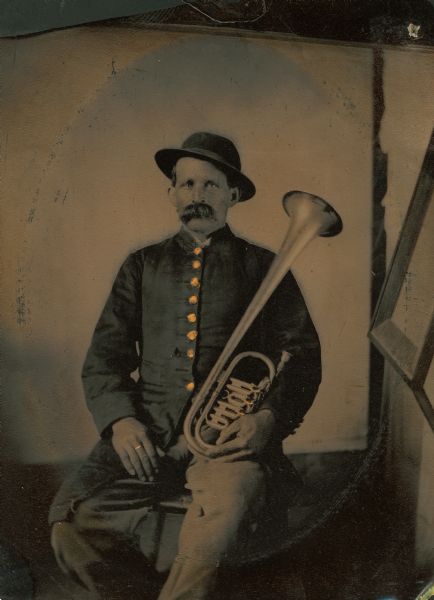 Tintype portrait of J.M. Faust of Broadhead, Wisconsin, member of the 3rd Wisconsin Infantry band. He is seated in uniform and holds an E-flat horn. The buttons on his uniform are hand-colored gold.