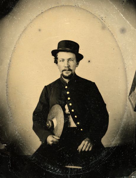 Tintype portrait of Norman Hall pf Brodhead, Wisconsin, member of the 3rd Wisconsin Infantry band. He is sitting and is wearing a uniform, and is holding a cymbal. The buttons on his uniform are hand-colored gold.