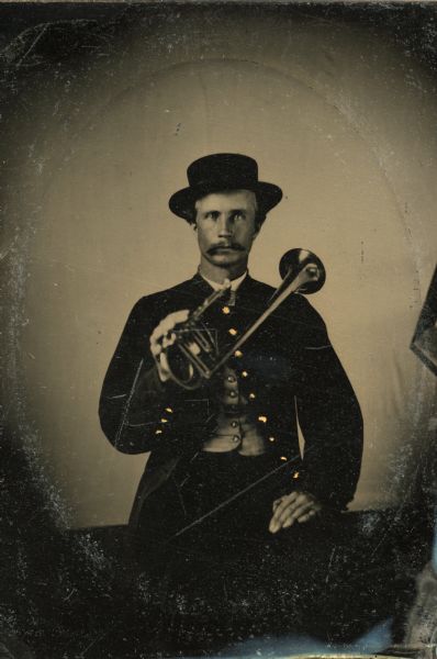 Tintype portrait of E.G. Kheeland of Monroe, Wisconsin, member of the 3rd Wisconsin Infantry band. He is sitting, and is wearing a uniform, and is holding a B-flat horn. The buttons on his uniform are hand-colored gold.