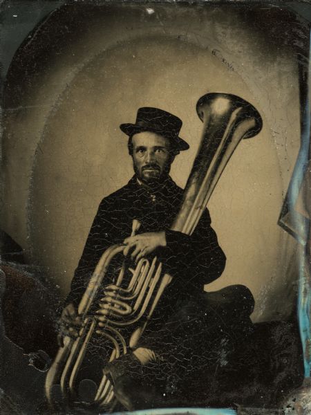 Tintype portrait of William Mason of Magnolia, Wisconsin, member of the 3rd Wisconsin Infantry band. He is seated in uniform and holds a tuba. The buttons on his uniform are hand-colored gold.