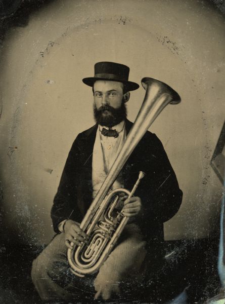 Tintype portrait of Theodore Pomeroy of Brodhead, Wisconsin, member of the 3rd Wisconsin Infantry band. He is seated in a suit and holds a tenor horn. The pin on his cravat is hand-colored gold.