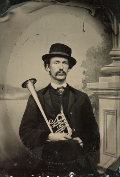 Tintype studio portrait in front of a painted background of George Spaulding of Brodhead, Wisconsin, member of the 3rd Wisconsin Infantry band. He is seated wearing a suit and holds a B-flat horn.