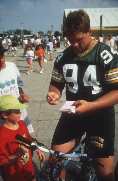 Green Bay Packer player (possibly Bob Kuberski) sitting on a young boy's bicycle and signing a notebook in the parking lot of Lambeau Field.